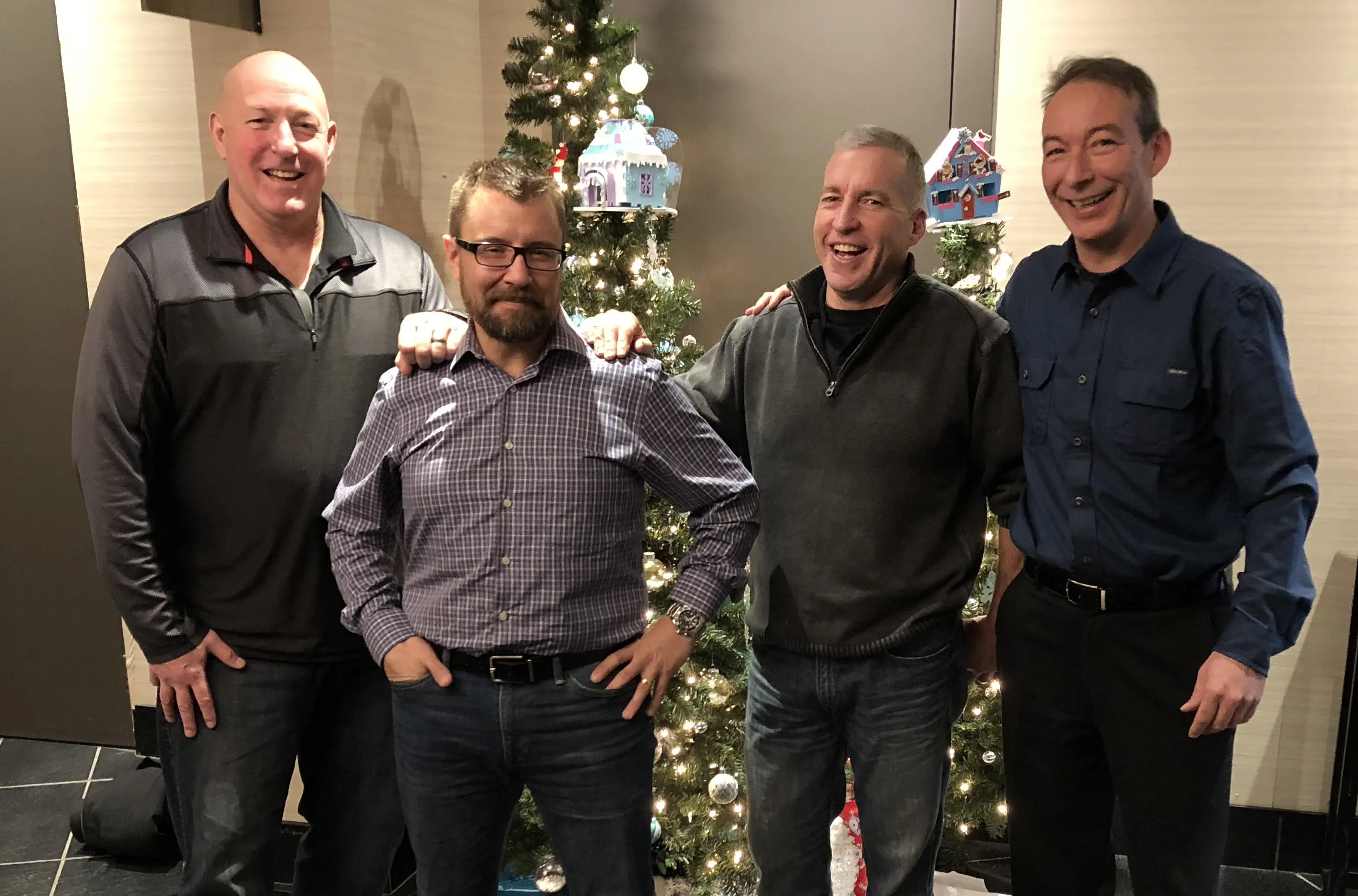 Four men standing and smiling in front of a decorated Christmas tree.